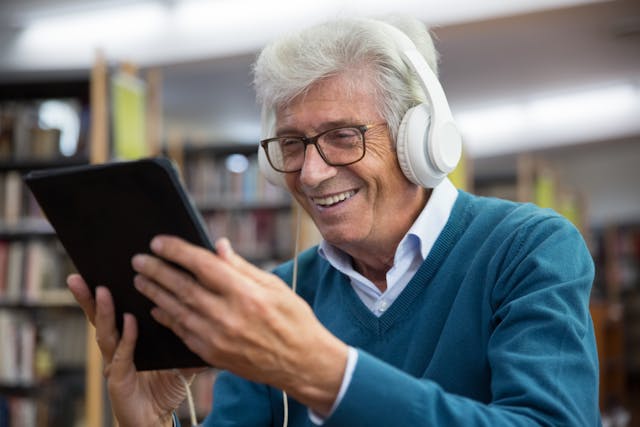older man sat in a library with headphones on using an ipad