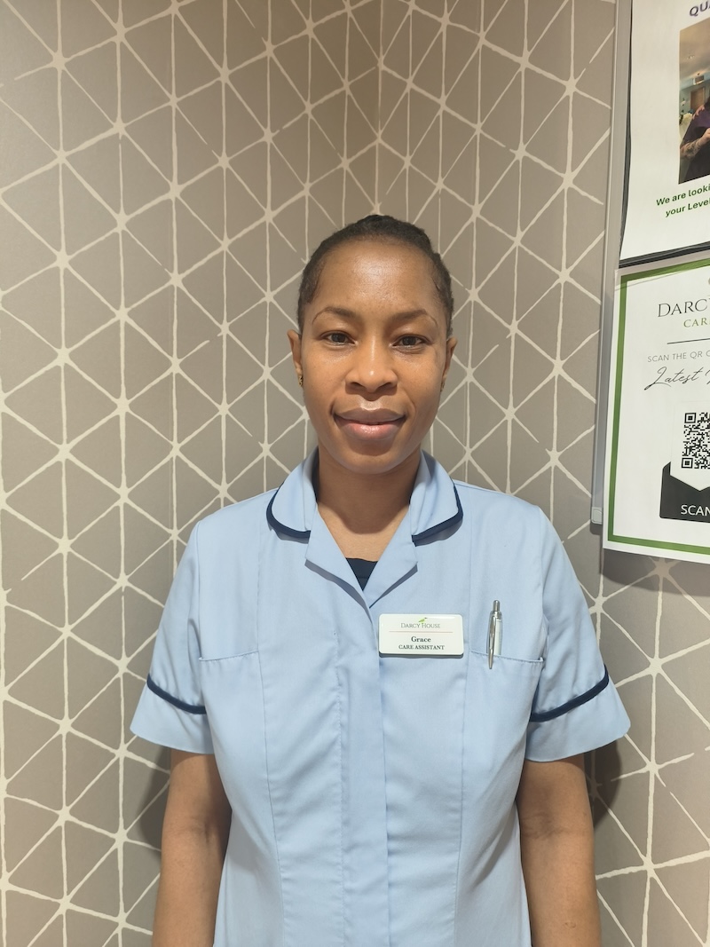 woman with dark hair tied back wearing a blue carers uniform having a headshot taken for the team page