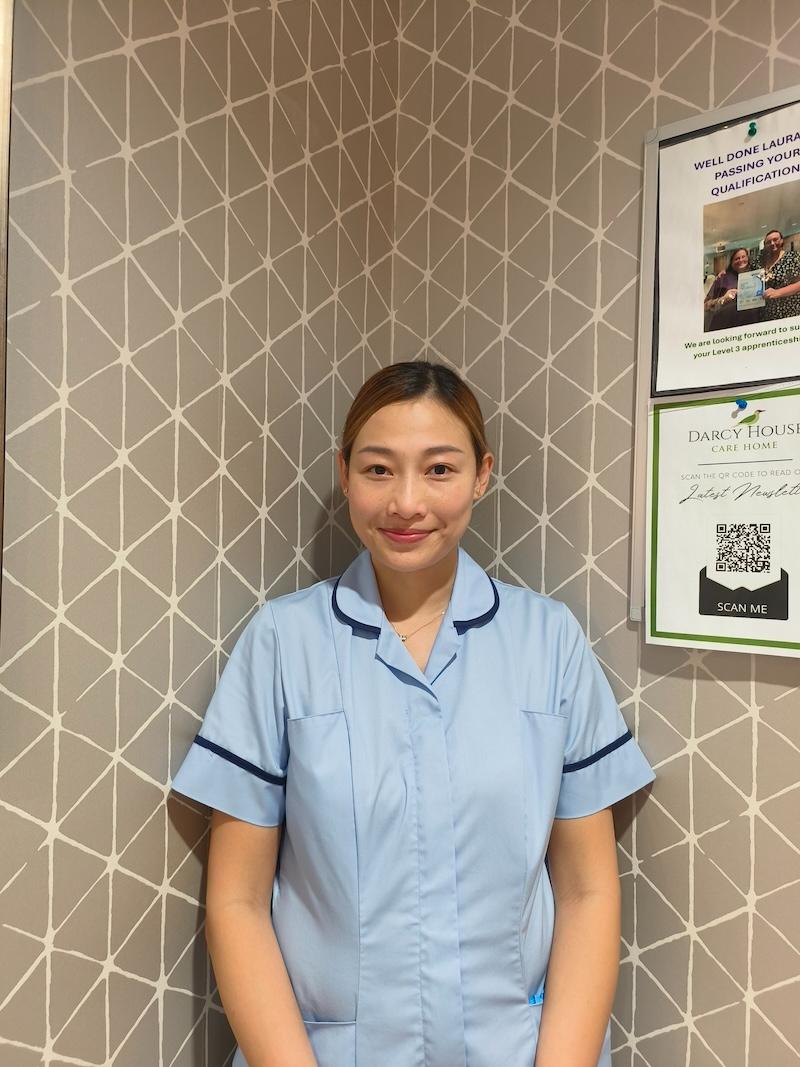 woman with brown hair tied back wearing a blue carers uniform having a headshot taken for the team page