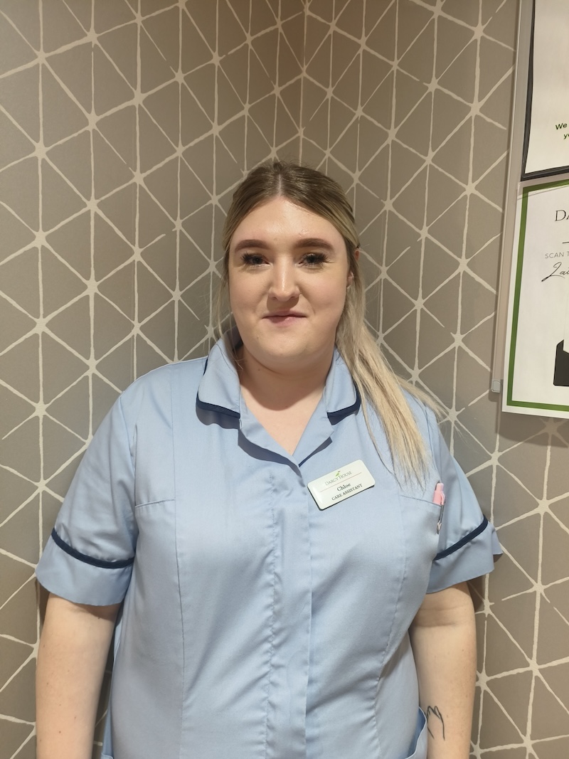 woman with blonde hair tied in a ponytail wearing a blue carers uniform having a headshot taken for the team page