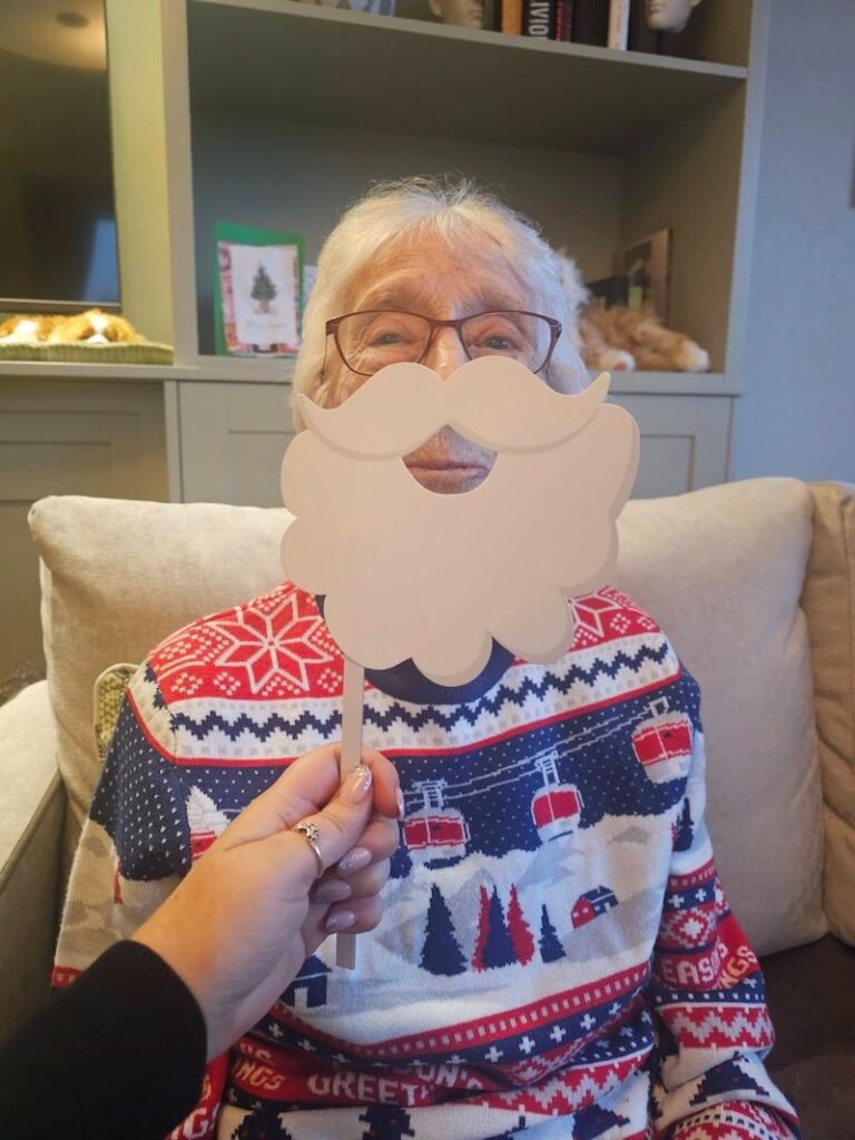 elderly resident sat in an armchair wearing a red, blue and white christmas jumper with a cardboard beard held up to their face